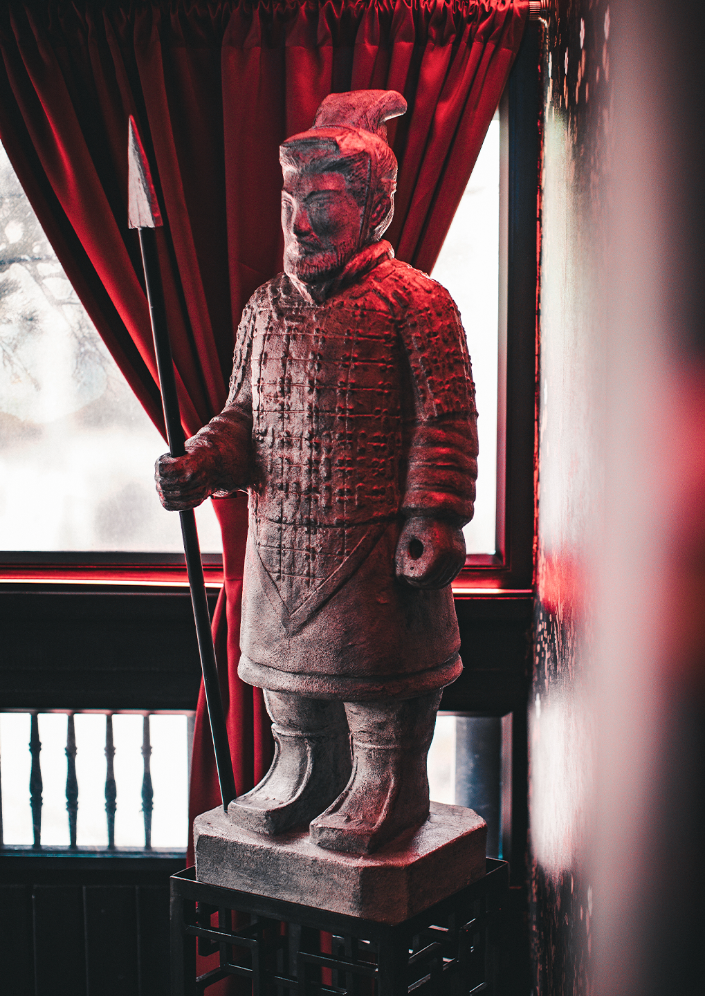 Terracotta warrior statue holding a spear, standing on a pedestal by a window with red curtains in a dimly lit room
