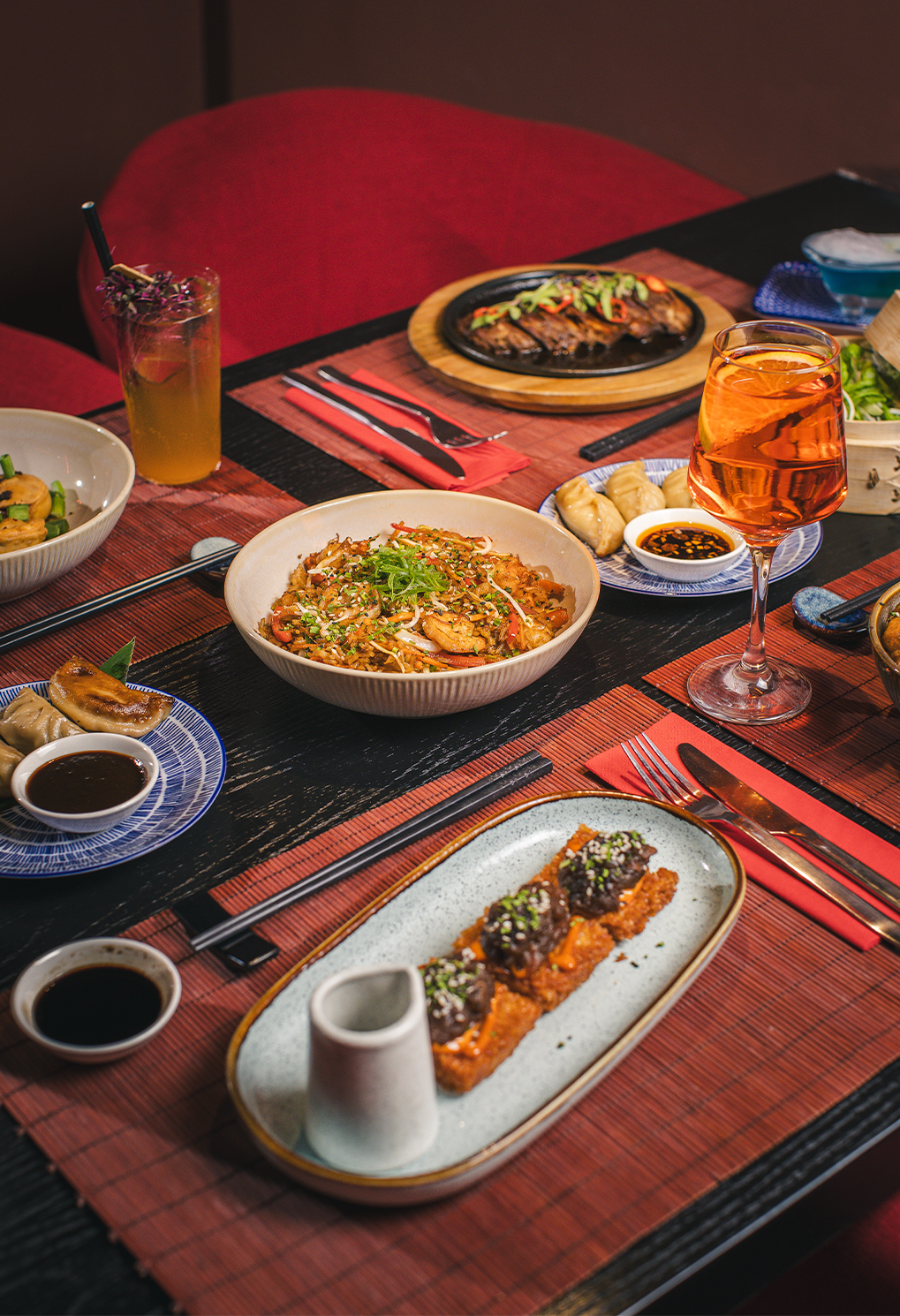 A table full of Asian dishes: a rice bowl centerpiece, dumplings with dipping sauce, sushi rolls, and a wine glass on a red placemat table setting.