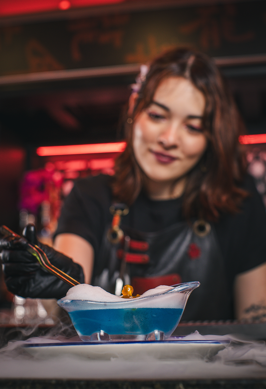 Female bartender adds an orange garnish to a blue foamy cocktail with dry-ice fog in a neon-lit bar setting.