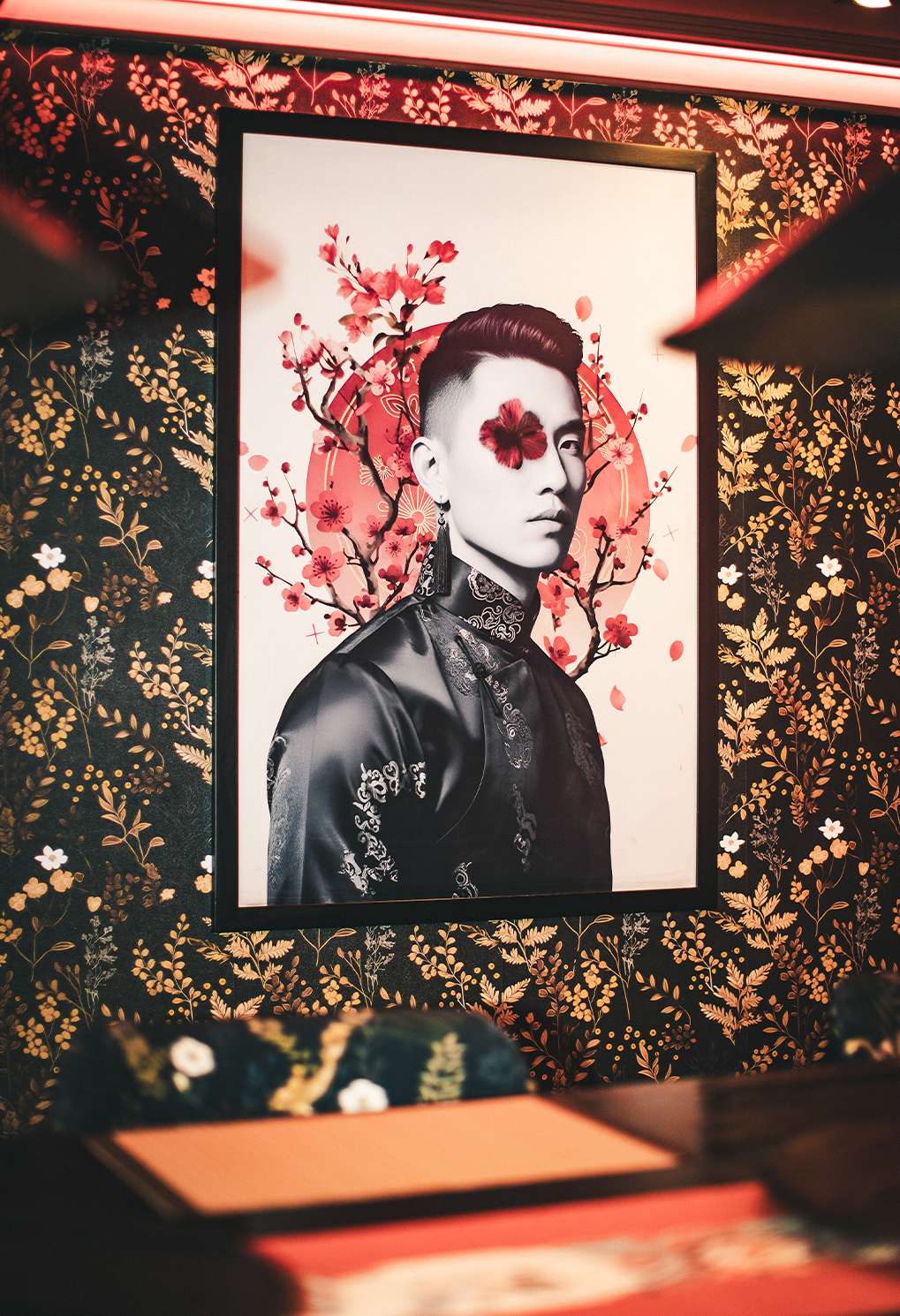 Framed black-and-white portrait of a person in a traditional embroidered outfit, with a red flower over one eye, displayed against floral wallpaper.