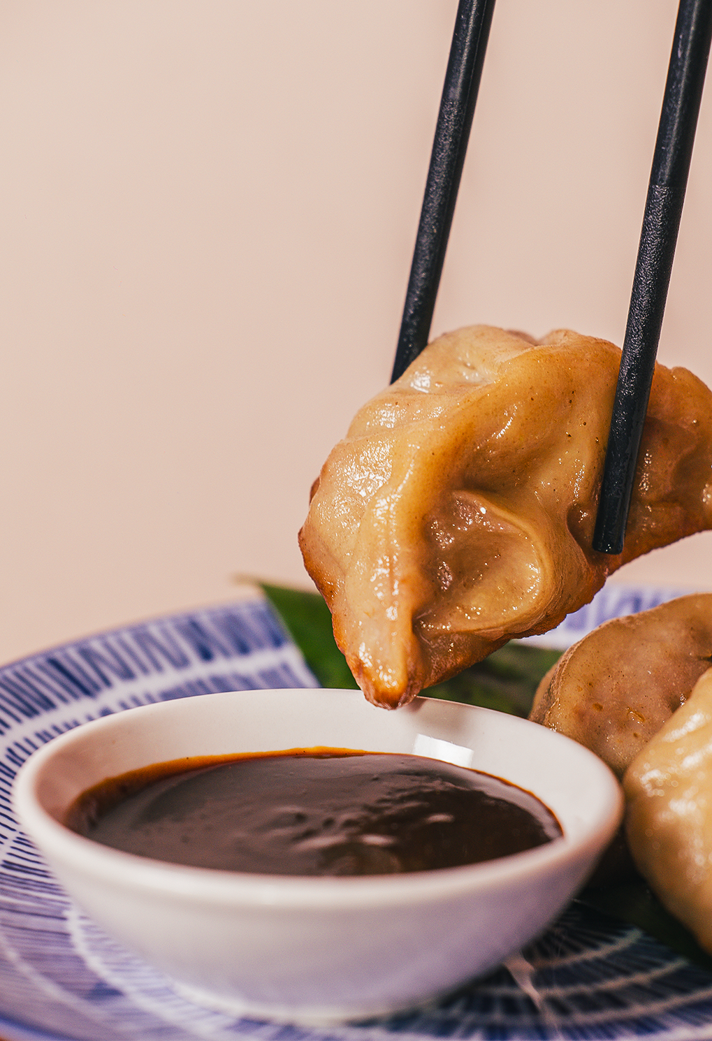 Dumpling being picked up with chopsticks over a white bowl of dark dipping sauce on a blue-patterned plate.