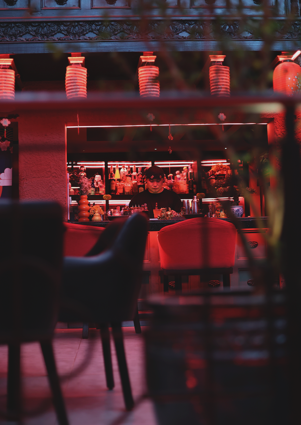 Bartender behind a neon red-lit bar with bottles on shelves and red chairs in the foreground.