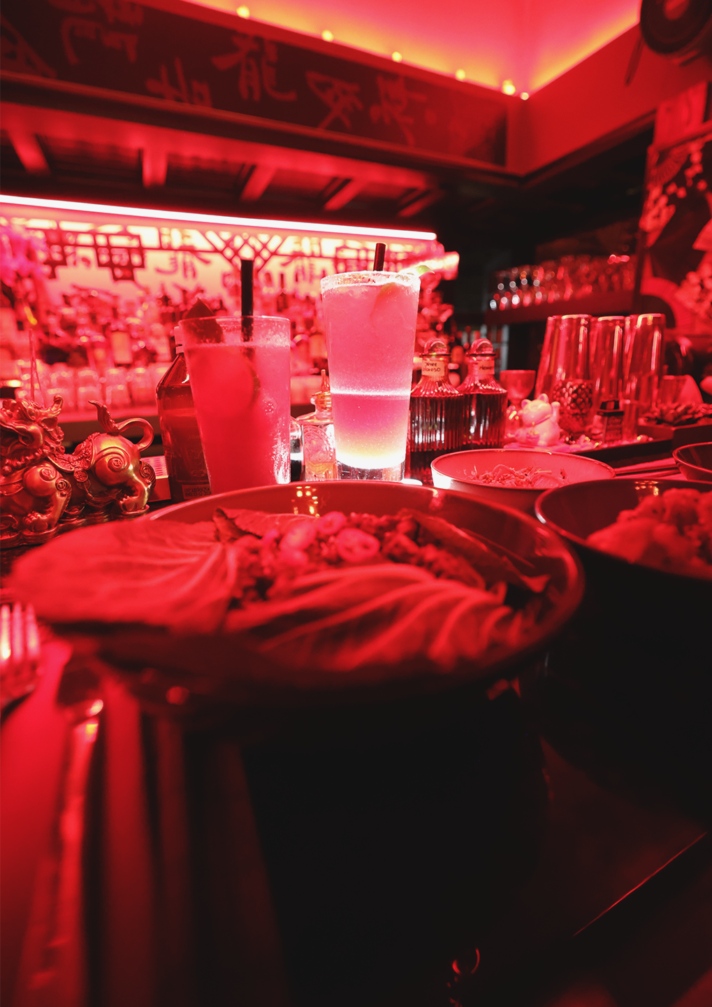 A red-lit bar scene with two frosted cocktails at the foreground and plates of food on the counter behind them.