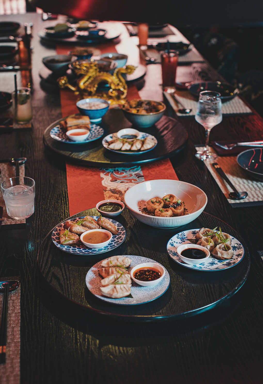 Bottom-front round dining table with plates of dumplings and dipping sauces, plus more dishes and drinks visible along a long wood table.