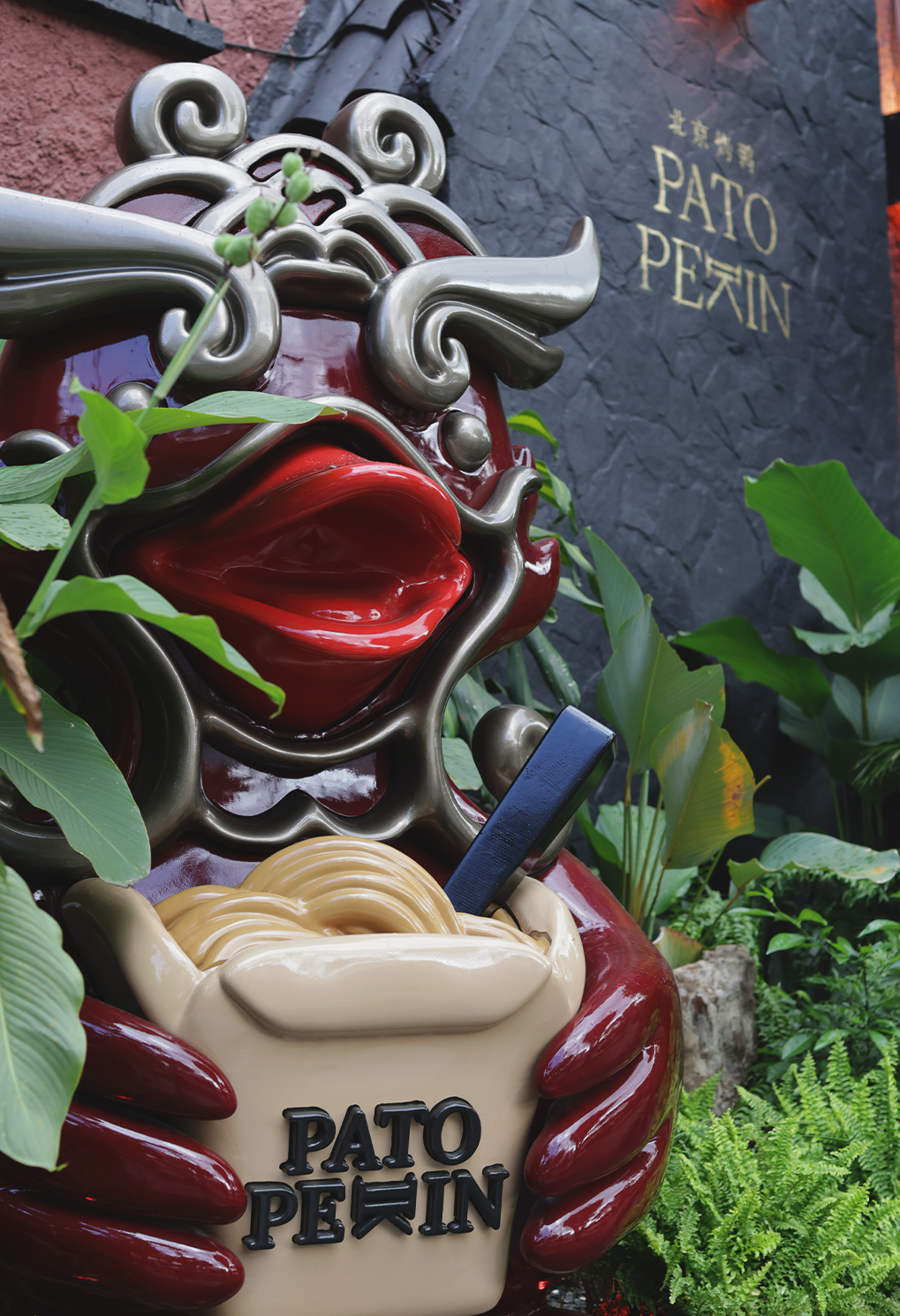 Decorative glossy sculpture of a stylized red figure with metal scrollwork, holding a beige purse labeled 'PATO PEKIN' amid green plants.