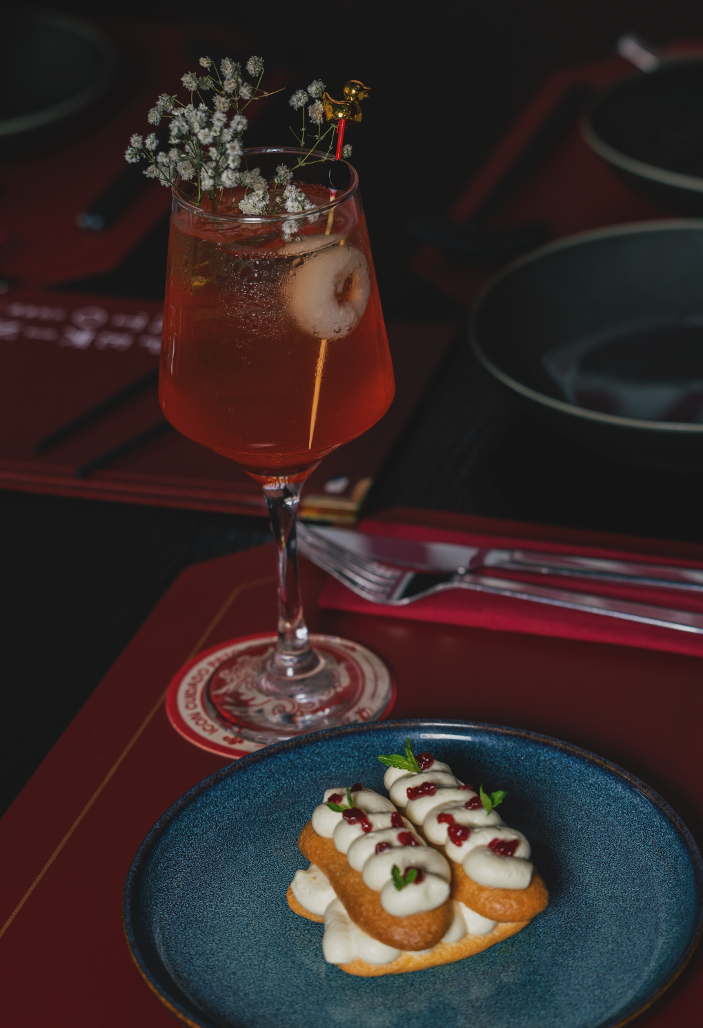 Red cocktail in a tall glass with ice, garnished with white baby's-breath flowers and a tiny gold figurine, plus two cream-topped éclairs on a blue plate nearby.