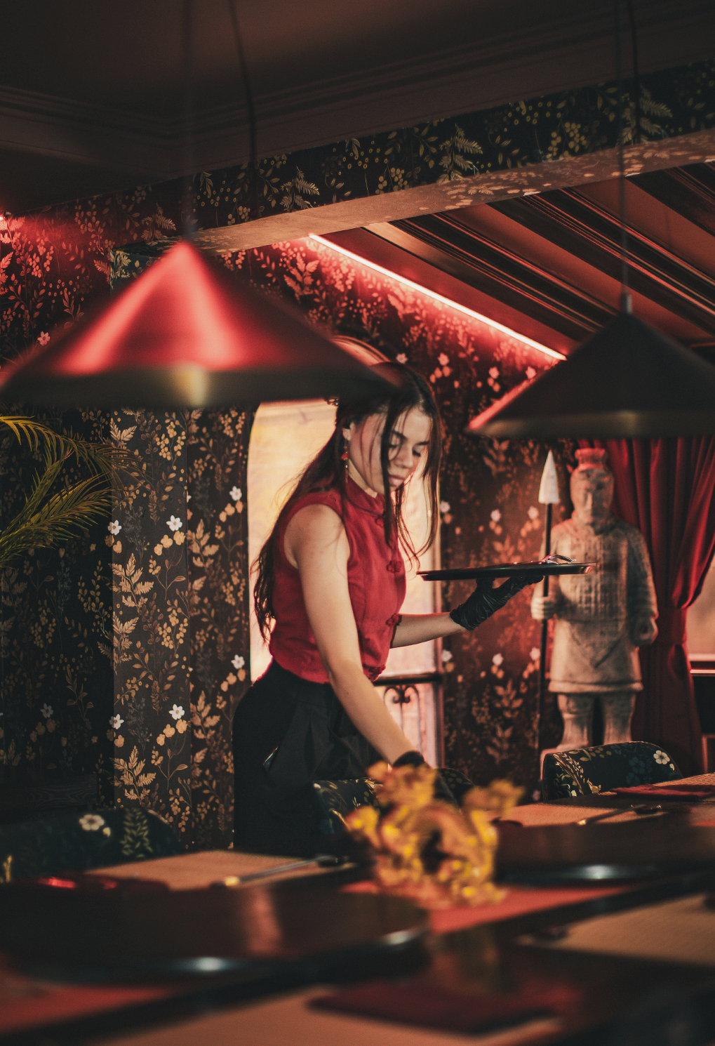 A young waitress in a red sleeveless top carries a tray in a dimly lit dining room with floral wallpaper patterns.