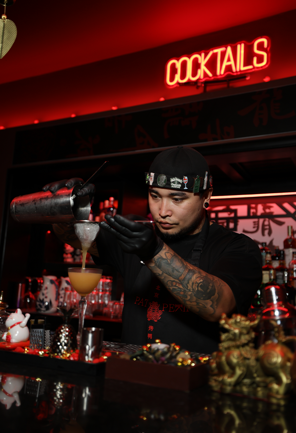 Bartender in black pours a cocktail from a shaker into a coupe glass at a red-lit bar with a neon COCKTAILS sign overhead and tattoos on his arm.