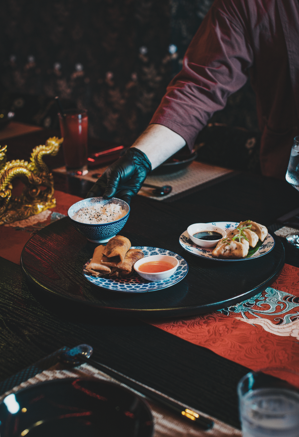 A server wearing a glove places dipping sauce and fried rolls on a blue patterned plate at a dark wooden table set with Asian-inspired decor,