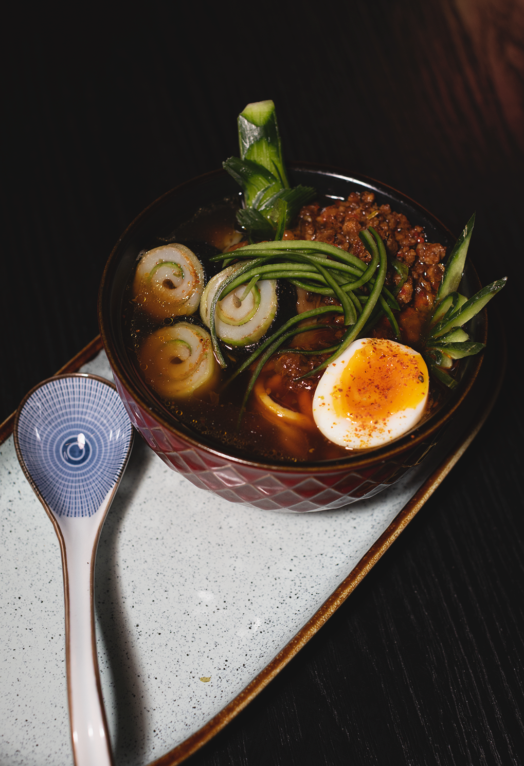 Ramen bowl with soft-boiled egg, minced meat, scallions, and greens in a dark broth.