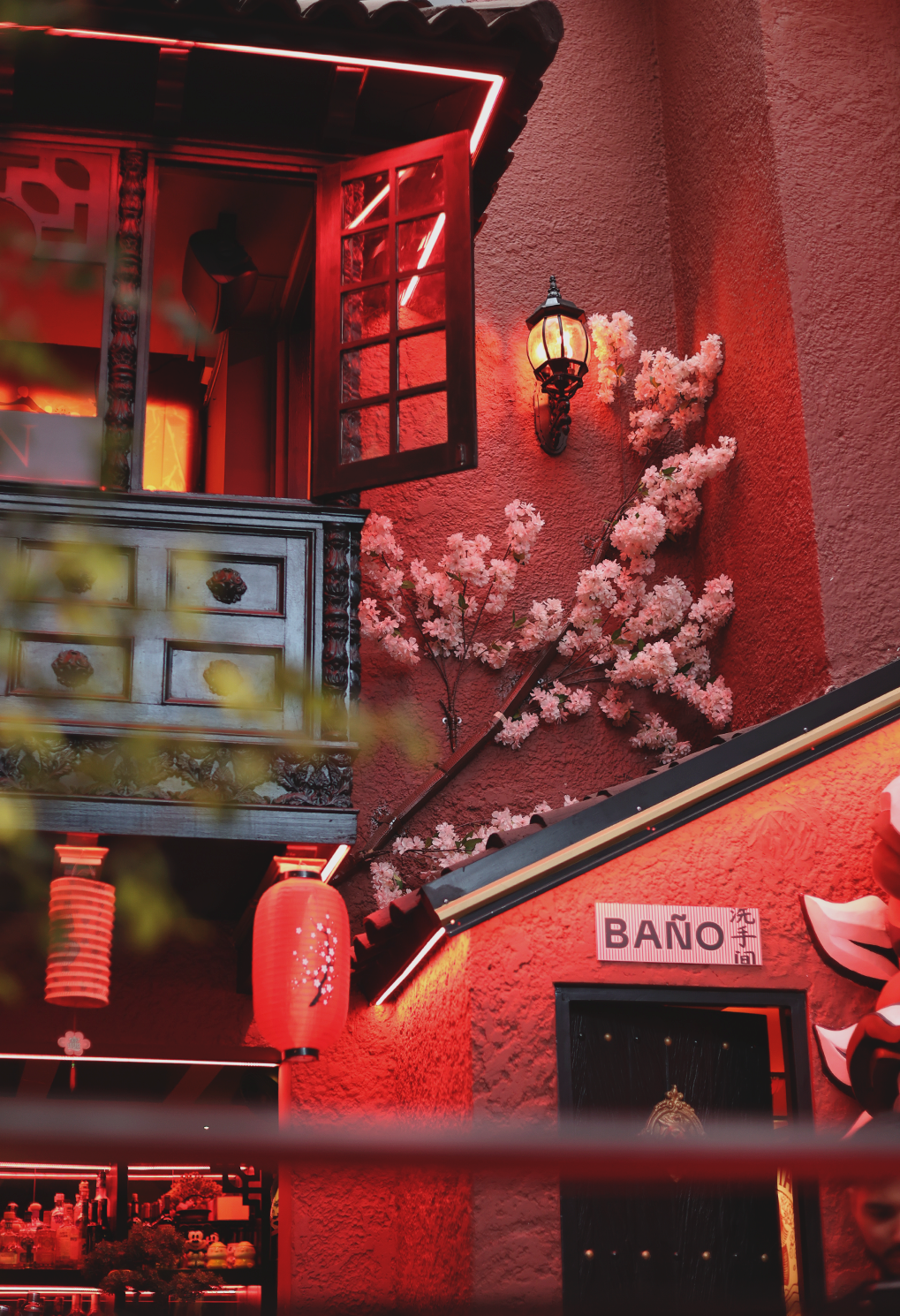 Red exterior of a cafe or shop with pink blossoms, red lanterns, and a glowing lantern on the wall under neon lighting.