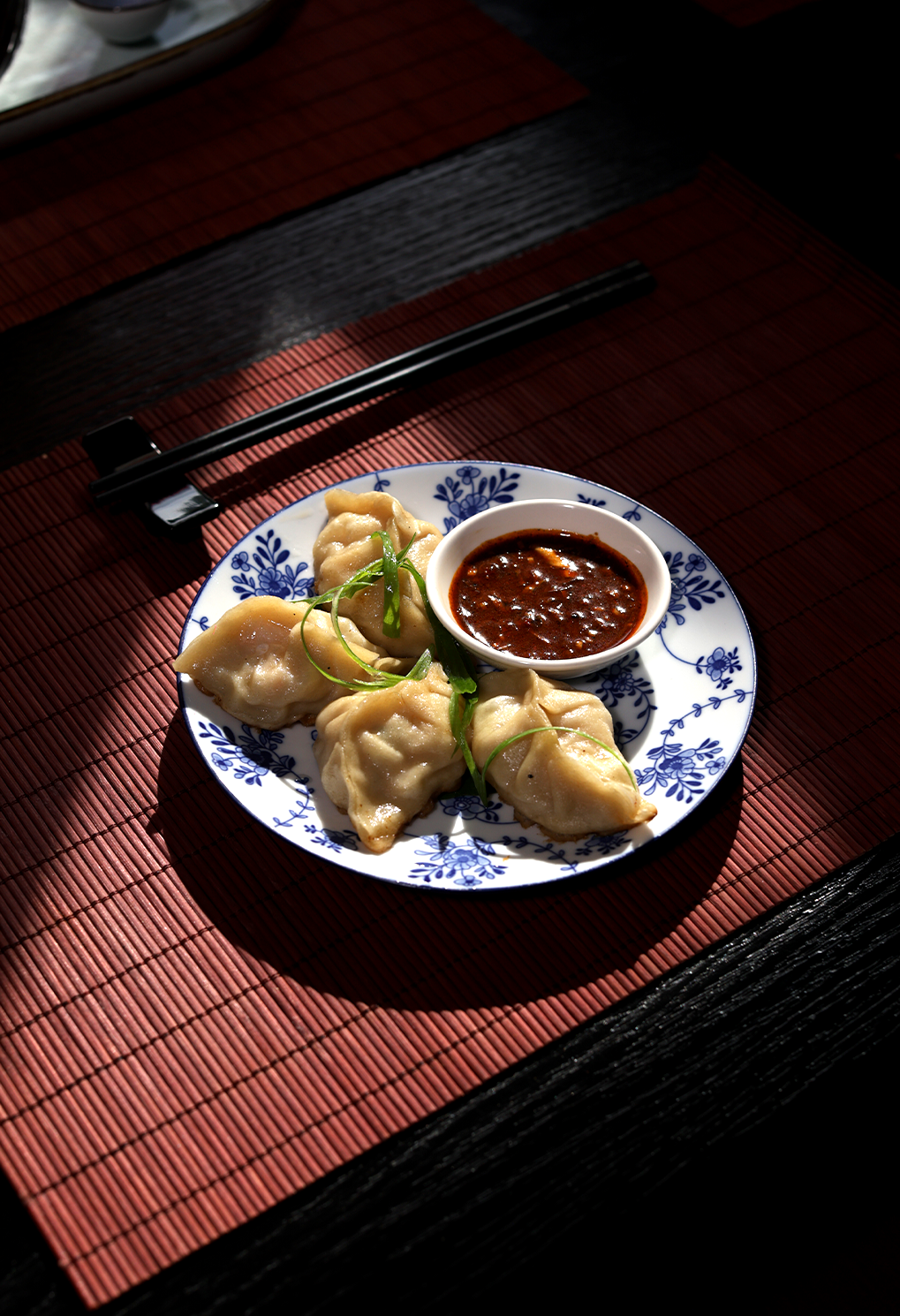 Dumplings arranged on a blue‑and‑white floral plate with a small bowl of dipping sauce, on a red placemat with chopsticks nearby.