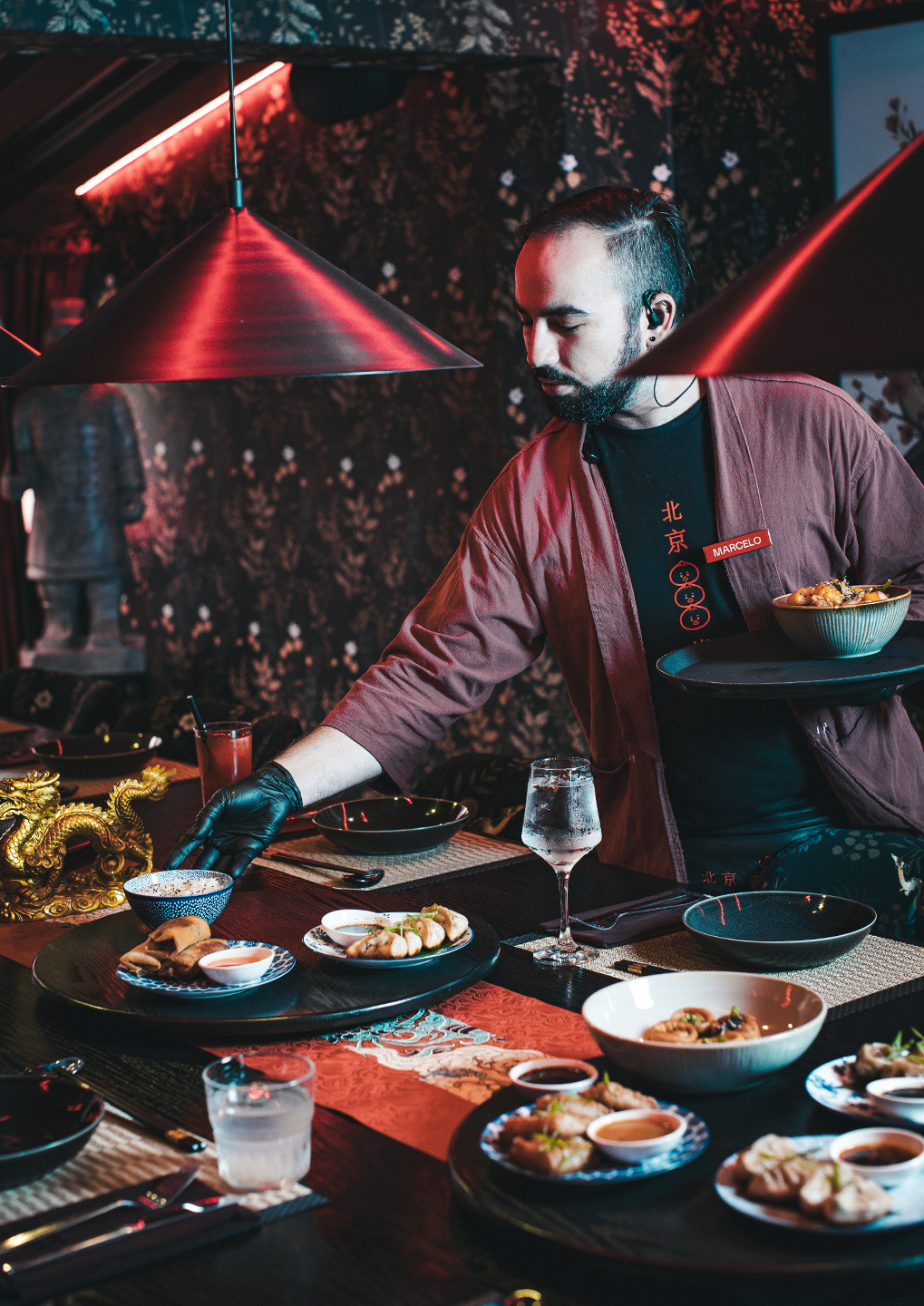 Bearded server in a reddish robe with a name tag, serving a bowl at a dark dining table under red pendant lights.