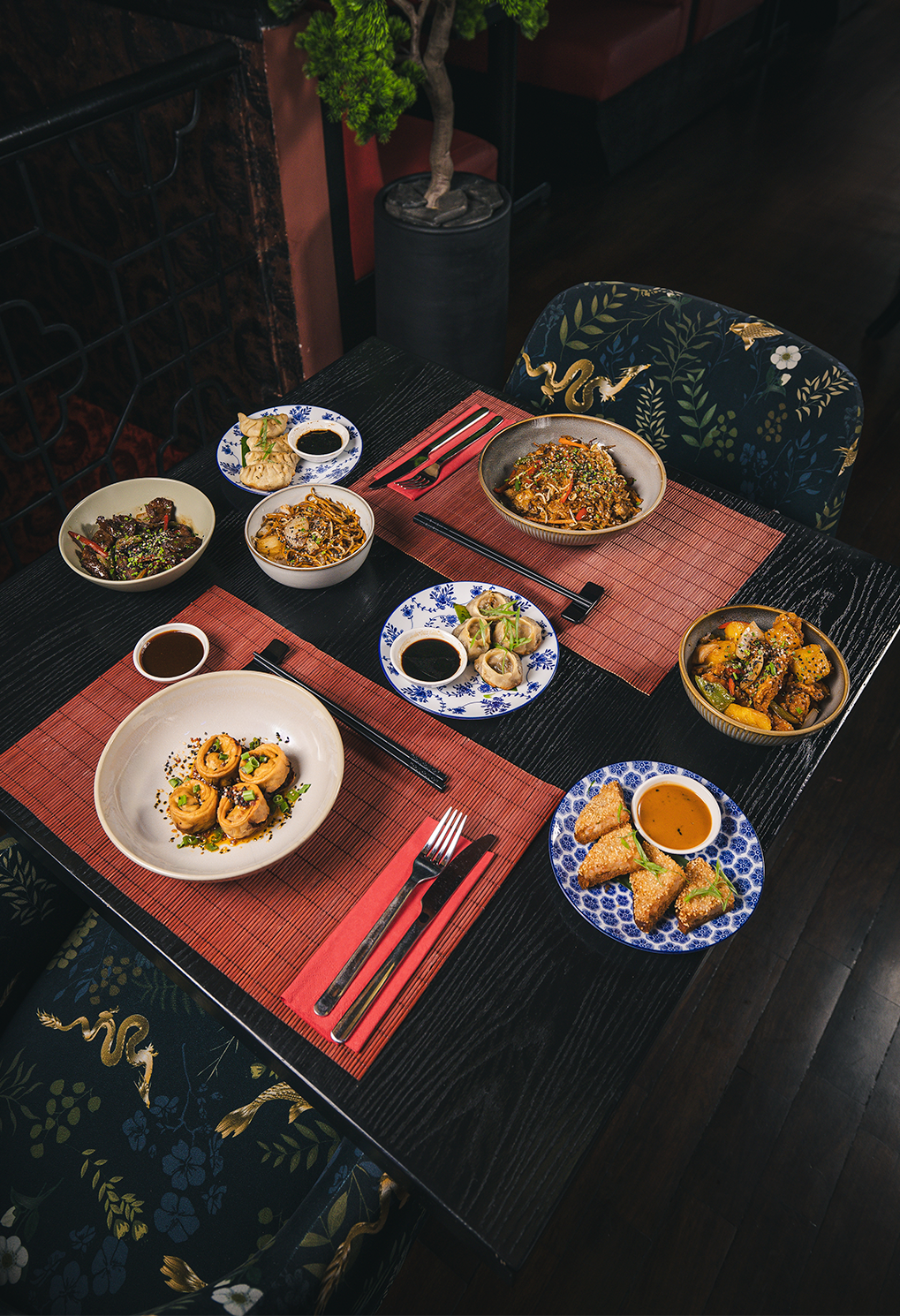 Overhead view of a dining table with multiple Asian dishes, dipping sauces, and chopsticks on red placemats.