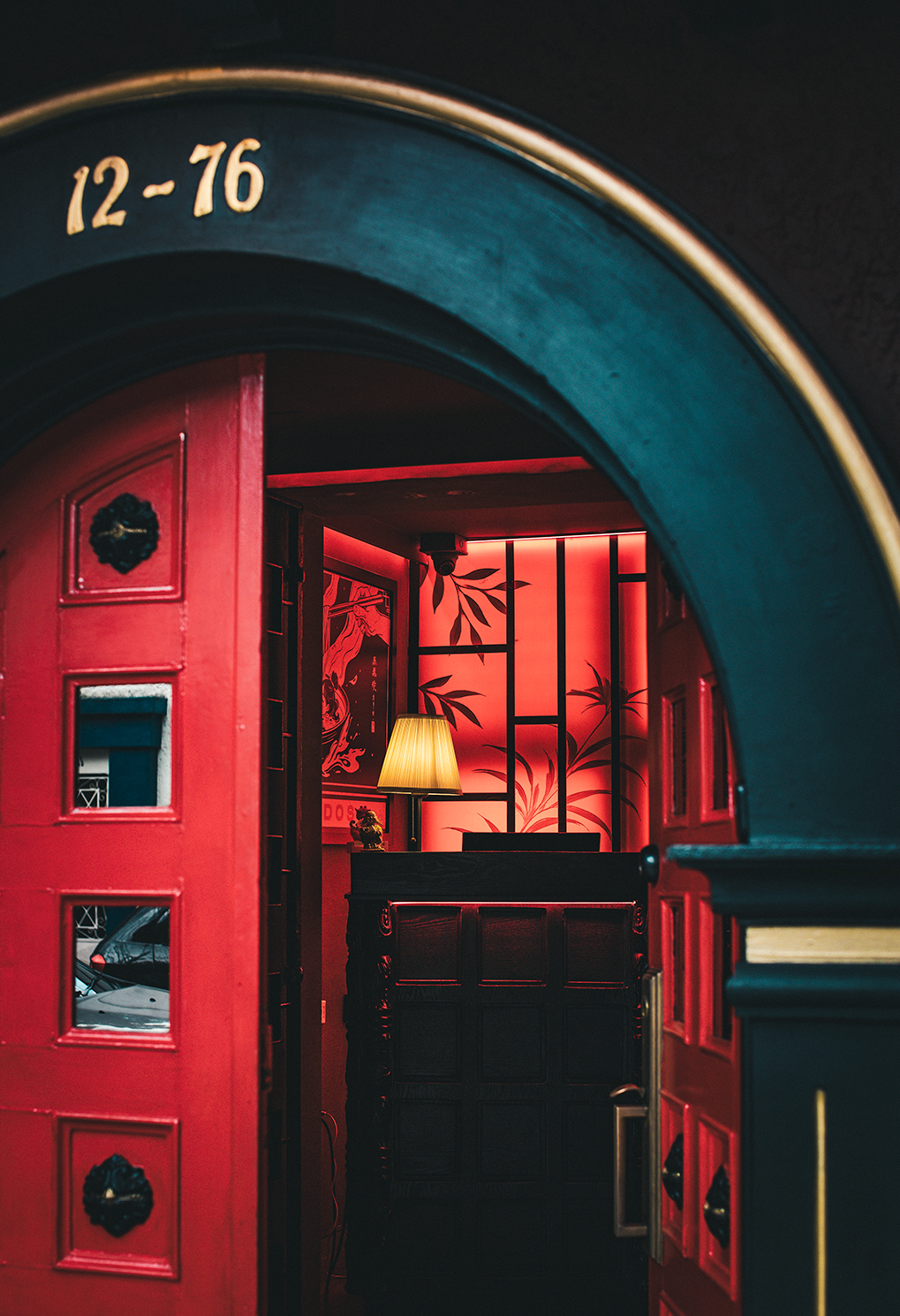 Red doorway opens to a warmly lit interior with a dark wooden counter and a lampshade against red bamboo-pattern wallpaper.