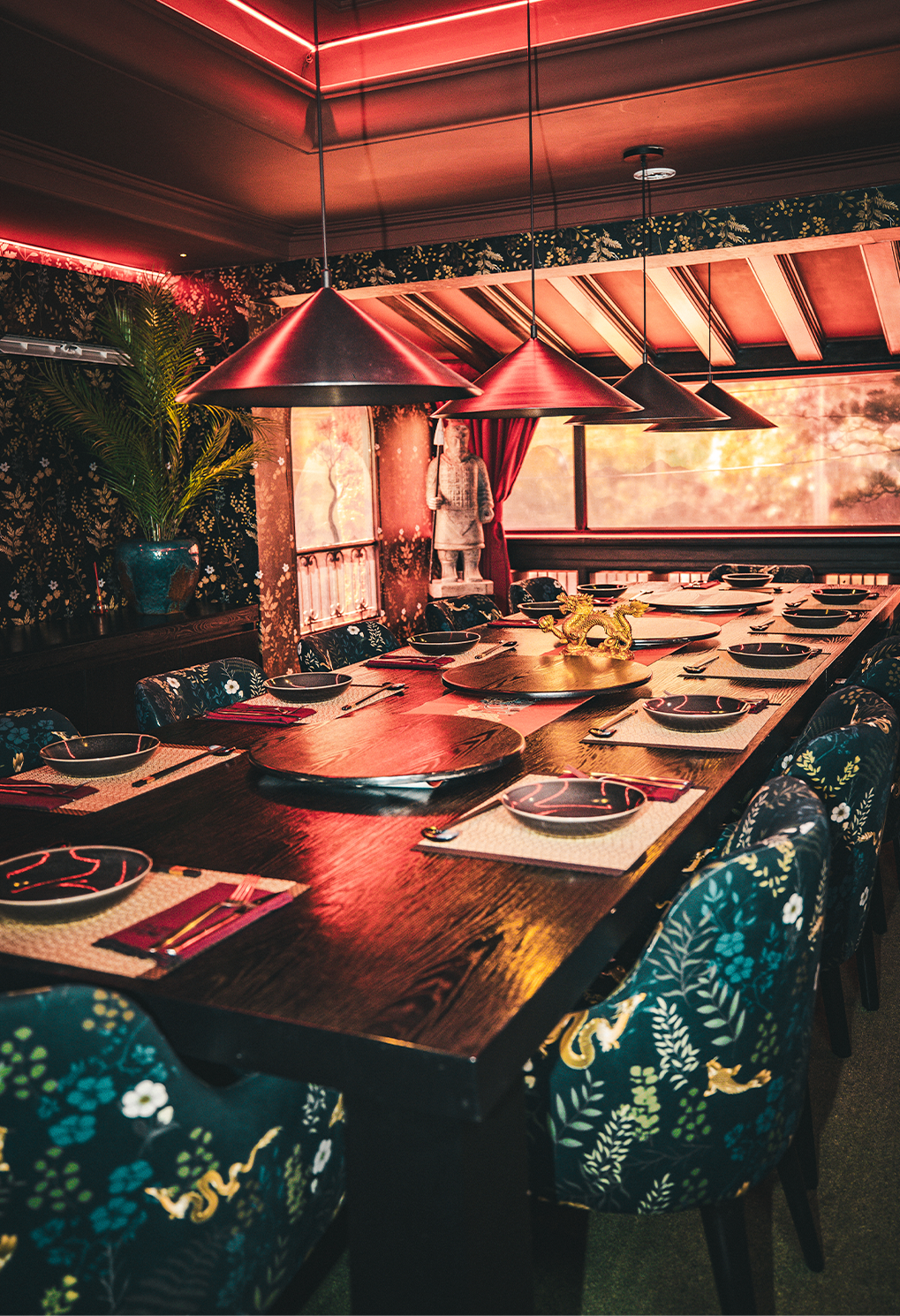 Long wooden dining table set for a group under red pendant lamps in a vintage floral restaurant interior with patterned chairs and a gold dragon centerpiece on a lazy Susan.