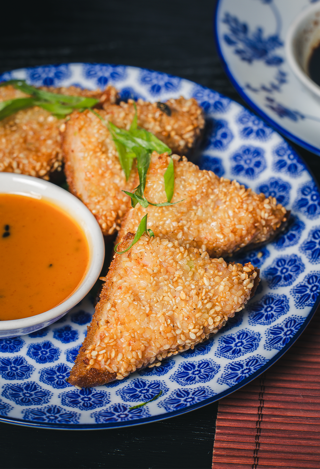 Sesame-crusted chicken wings with green garnish on a blue patterned plate and orange dipping sauce.