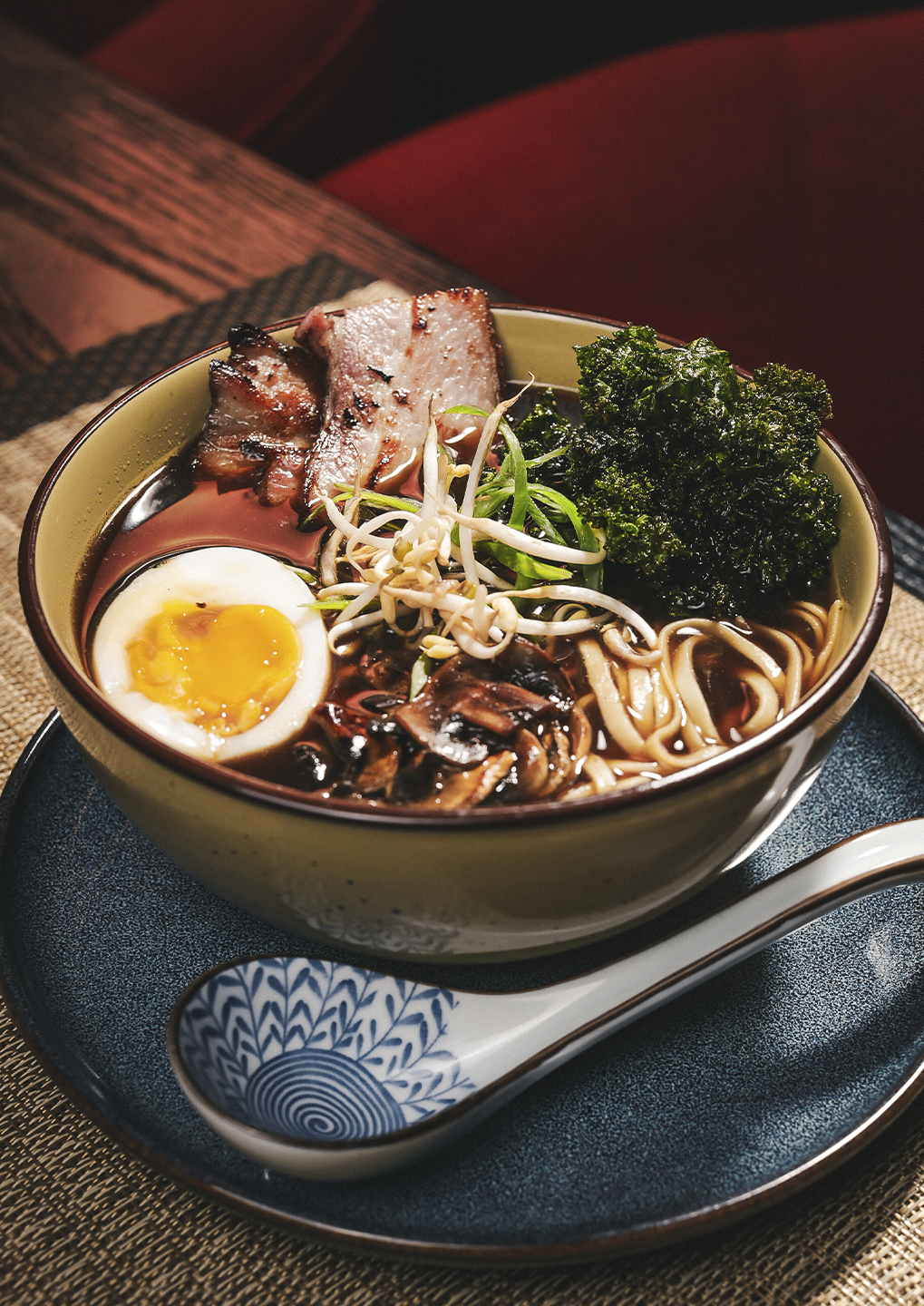 Bowl of ramen with sliced pork, a soft-boiled egg, bean sprouts, greens, and noodles in broth on a blue plate