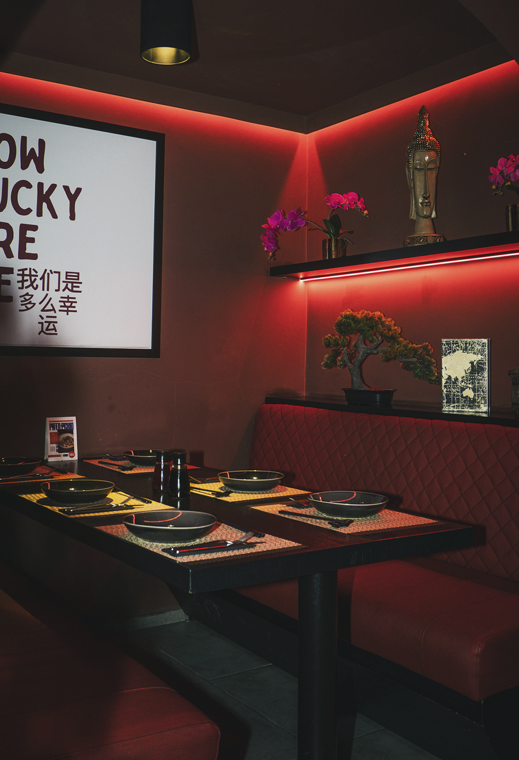 Cozy restaurant booth with a red upholstered banquette, a black table set with bowls and place settings under red ambient lighting.
