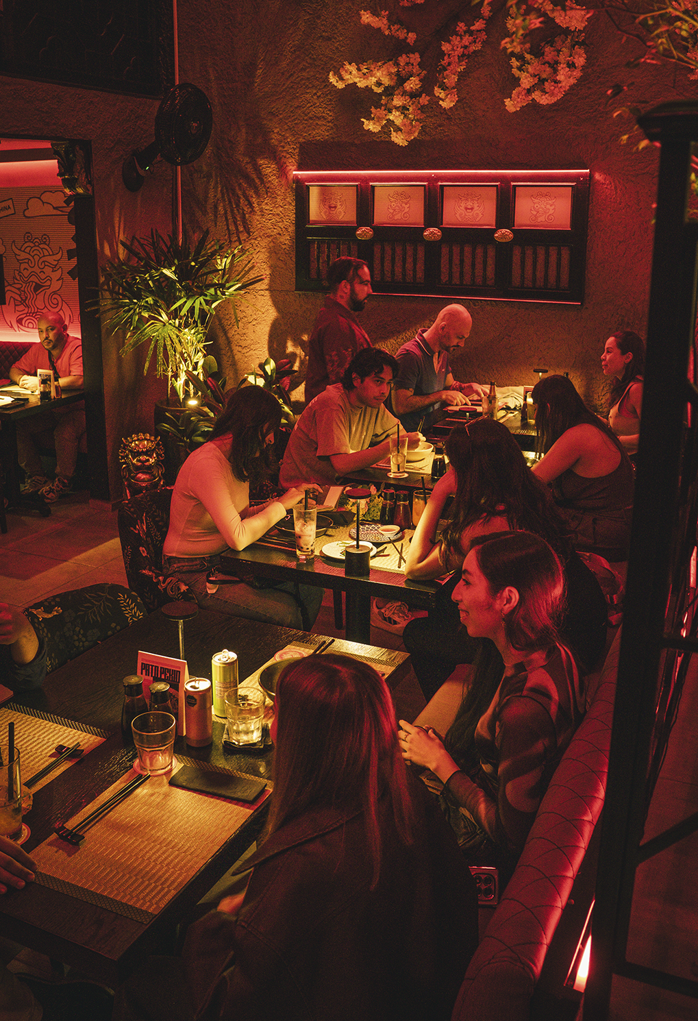 People dining at tables in a cozy, red-lit restaurant with plants and wall art in the background.