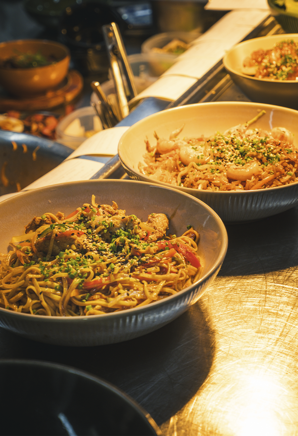 Two bowls of stir-fried noodles with vegetables and sesame seeds on a stainless steel counter in a kitchen setting.