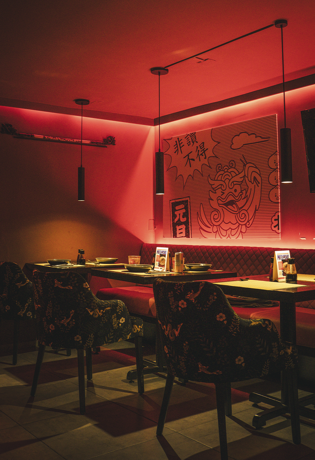 Restaurant dining area with red ambient lighting, booth seating, and tables set for service under hanging pendant lights. The wall features bold artwork with a dragon motif and Asian characters.